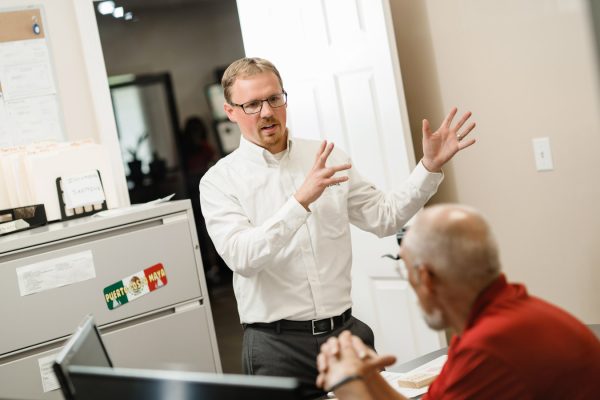 A male professional in Information Security Discusses Cybersecurity Risks in an Office Environment. The trainer has his hands demonstrating something, and is facing a user and photographer. The user has hands folded and is facing the trainer.